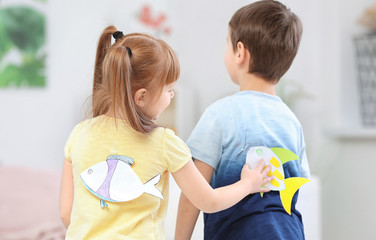 Little girl sticking paper fish to boy's back indoors. April fool's day prank