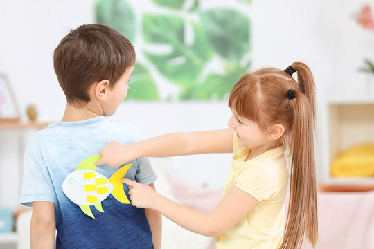 Little girl sticking paper fish to boy's back indoors. April fool's day prank