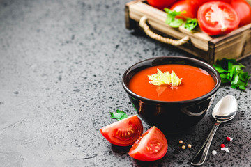 Red pepper tomato soup in a bowl, fresh tomatoes in a wooden box on dark concrete background. Selective focus, space for text.