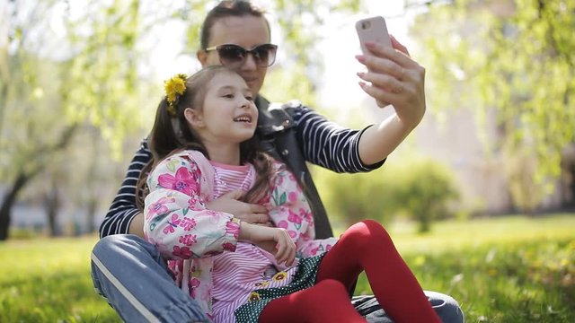 Photo selfie family - mother and little child daughter hugging kissing shooting pictures via smart phone during walking in park