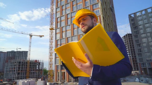 young attractive man in a yellow hard hat looks into the camera. Portrait of a handsome business man. the builder makes notes in a yellow folder. high-rise buildings under construction in the