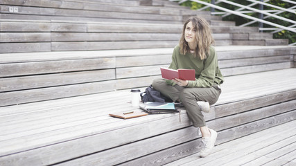 Fototapeta na wymiar A young brunette girl dressed in a green sweat shirt is sitting on the stairs holding a book on the stairs looking to the sky