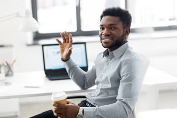 business and people concept - african american businessman with coffee sitting at office table and waving hand