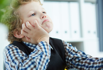 Tired little boy propped his head in his hands and looks at the monitor at the office