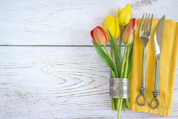 Festive table setting with original cutlery on yellow napkin and bouquet of tulips in napkin ring on white wooden background with copy space