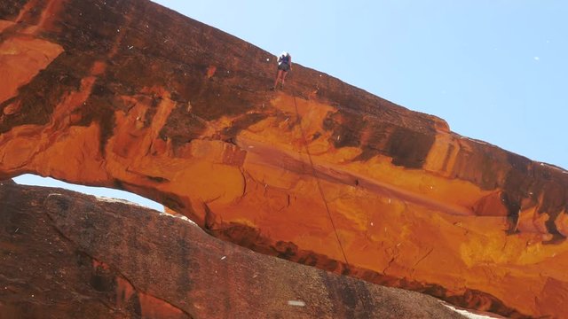 A red rock arch or land bridge carved out of sandstone. A adventurous woman repels down to the safe ground.