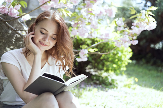 Young beautiful redhead girl in white t-shirt and gray skirt reads a book for her blogg channel in sunny springtime blossom cherry trees garden. Hobby, vlogger, blogger, analog photo, social media. 