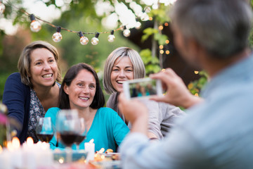 One summer evening friends gathered around a table in the garden for a good time. A man takes a picture of three female friends in their forties