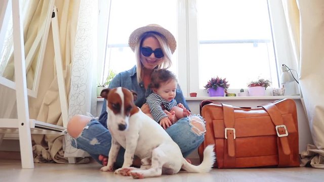 Young mother with a child sitting on a floor near dog and suitcase with a window. Person is ready for summertime vacation