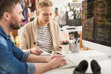 Portrait of creative young man writing code sitting at desk using computer and collaborating with colleague on startup project in modern office