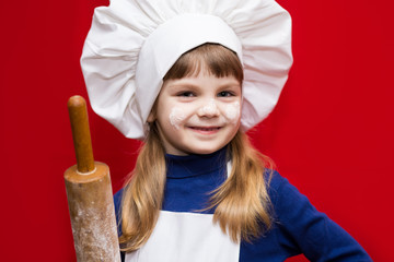 Happy little girl in chef uniform holds rolling pin isolated on red. Kid chef. Cooking Process Concept 