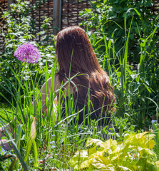 Junge rothaarige Frau mit langem Haar sitzt mitten im Garten, von hinten, von Blattgrün umgeben