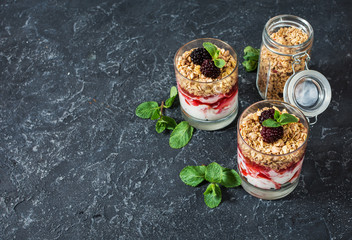 Healthy layered dessert with yogurt, granola, jam, blackberry in glass on stone background.