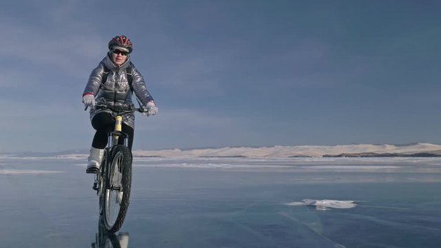 Woman is riding bicycle on the ice. The girl is dressed in a silvery down jacket, cycling backpack and helmet. Ice of the frozen Lake Baikal. The tires on the bicycle are covered with special spikes