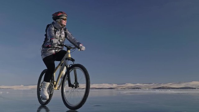 Woman is riding bicycle on the ice. The girl is dressed in a silvery down jacket, cycling backpack and helmet. Ice of the frozen Lake Baikal. The tires on the bicycle are covered with special spikes