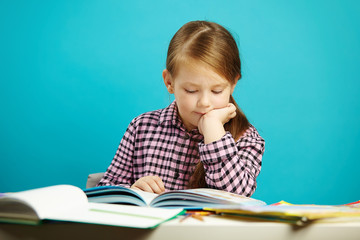 Disciplined girl attentively reads book sitting at the table, expresses her passion, on isolated blue background. Child performs home task.