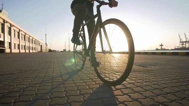 Stylish handsome man riding bicycle during sunset or sunrise with sea port on background, slow motion
