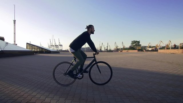 Stylish handsome man riding bicycle during sunset or sunrise with sea port on background