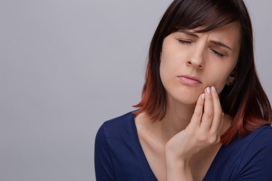 Closeup portrait of young woman on grey background suffering from toothache, holding fingers to jaw and closing eyes from pain.