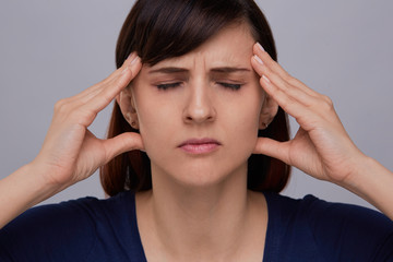 Closeup portrait of young woman on grey background suffering from strong headache, holding fingers to temples and closing eyes from pain.Severe migraine.