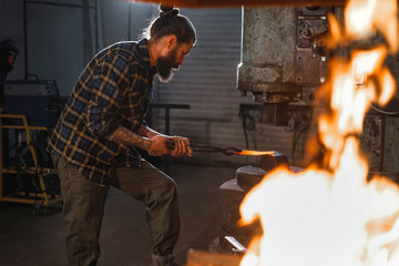 The smith works in his workshop on a mechanical hammer holding a hot piece in special tongs....