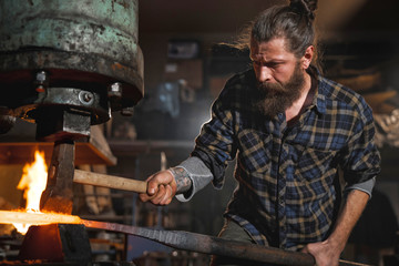 Real brutal blacksmith works in a workshop mechanical hammer with a red-hot iron. Portrait of a profession. - obrazy, fototapety, plakaty