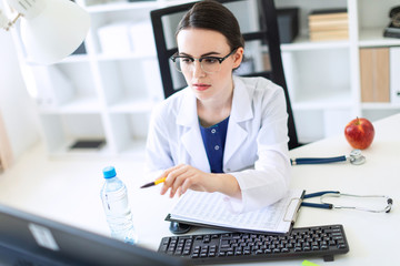 A beautiful young girl in a white robe is sitting at a computer desk with documents and a pen in her hands.