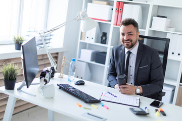 A man is sitting in the office at the table and holding a glass of coffee.