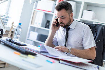 A man is sitting in the office, working with documents and talking on the phone.