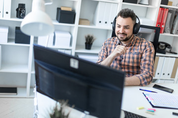 A young man in headphones sits at a table in the office and looks at the monitor.