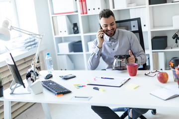 A young man in the office sits at a table, talking on the phone and holding a coffee pot.