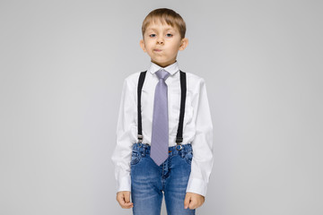 A charming boy in a white shirt, suspenders, a tie and light jeans stands on a gray background. the boy twisted his face