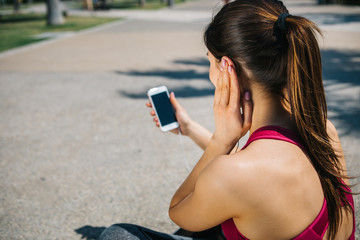 Back view of relaxed sportswoman is listening to music from earphones with enjoyment. She is holding a phone while sitting on alley in park. Copy space