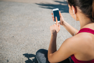 Cheerful young woman is relaxing after sport activity. She is touching smartphone screen while listening to music from earphones. Athlete is sitting on the road. Copy space