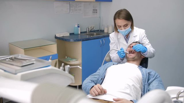 Front view of female dentist in gloves and face mask sitting behind male patient and treating his teeth with drill machine at modern dental clinic. Young patient receiving treatment for cavity