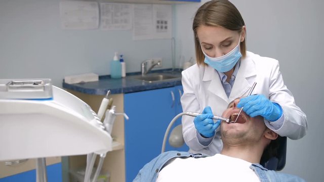 Portrait of attractive mid adult female dental doctor conducting tooth decay treatment with drill machine on a young male patient. Dentist in mask and gloves treating cavities at dental clinic