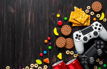 black and white modern gamepads on a dark wooden background among sweets, biscuits and cans of soda.