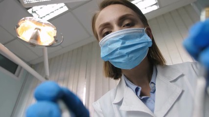 Attractive female mid adult dentist in white lab coat putting on protective face mask and starts examining client's teeth during dental check-up. Low angle shot, patient point of view