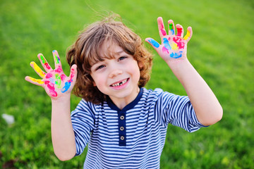 cheerful child boy with curly hair without front milk tooth shows hands dirty with multi-colored finger paints and smiles.