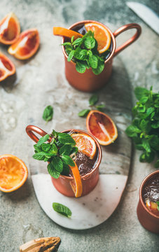 Blood orange Moscow mule alcohol cocktails with fresh mint leaves and ice in copper mugs on board in center over grey concrete background