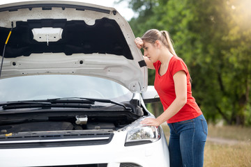 Portrait of sad young woman leaning on open car hood and looking at broken engine