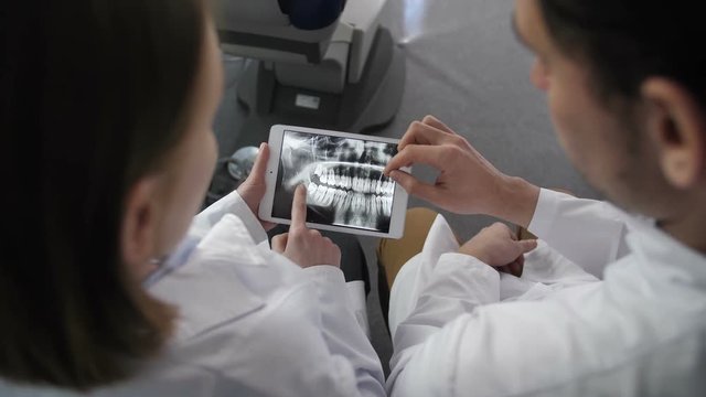 Tope angle view of young female and male dentists in laboratory coats using touchpad, looking at patient's dental x-ray on tablet pc at dental clinic. Medical dental doctors discussing diognosis