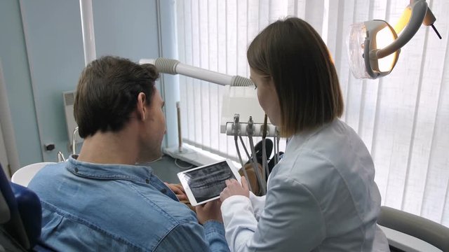 Back view of female dentist in white lab coat and young male patient in dental chair looking and talking about dental radiography x-ray at dental clinic. Doctor showing teeth x-ray on digital touchpad