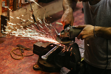 Blacksmith polishes crossguard of sword. Man is working in workshop. He holds a grinder in his hands.