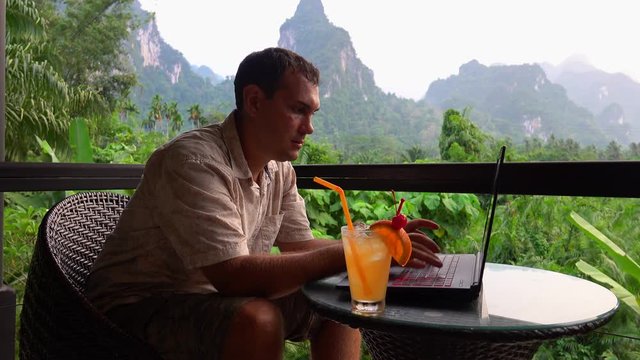 Male Traveler Using Laptop with Scenic Limestone Rocks View. Digital Nomad Working on Terace. Freelancer Typing on Computer in Asia. Remote Work in Thailand