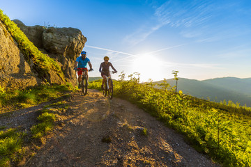 Fototapeta premium Cycling women and man riding on bikes at sunset mountains forest landscape. Couple cycling MTB enduro flow trail track. Outdoor sport activity.