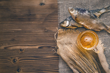 Foam beer in a mug, dried fish stockfish and rye ears wrapped in burlap cloth on a burnt wooden...