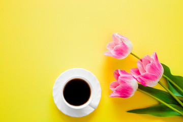 White cup of black coffee with pink tulips on bright yellow table. Coffee with flowers as lovely surprise from beloved. Positive morning. Empty place for inspirational, motivational text or quote.