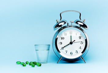Medical green capsule pills, glass water and alarm clock on blue background. Concept health