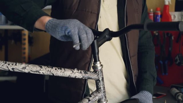 Tilt-down shot of young man professional repairman in earphones fixing bike handlebar using allen key and holding stearing wheel with hand in protective glove.